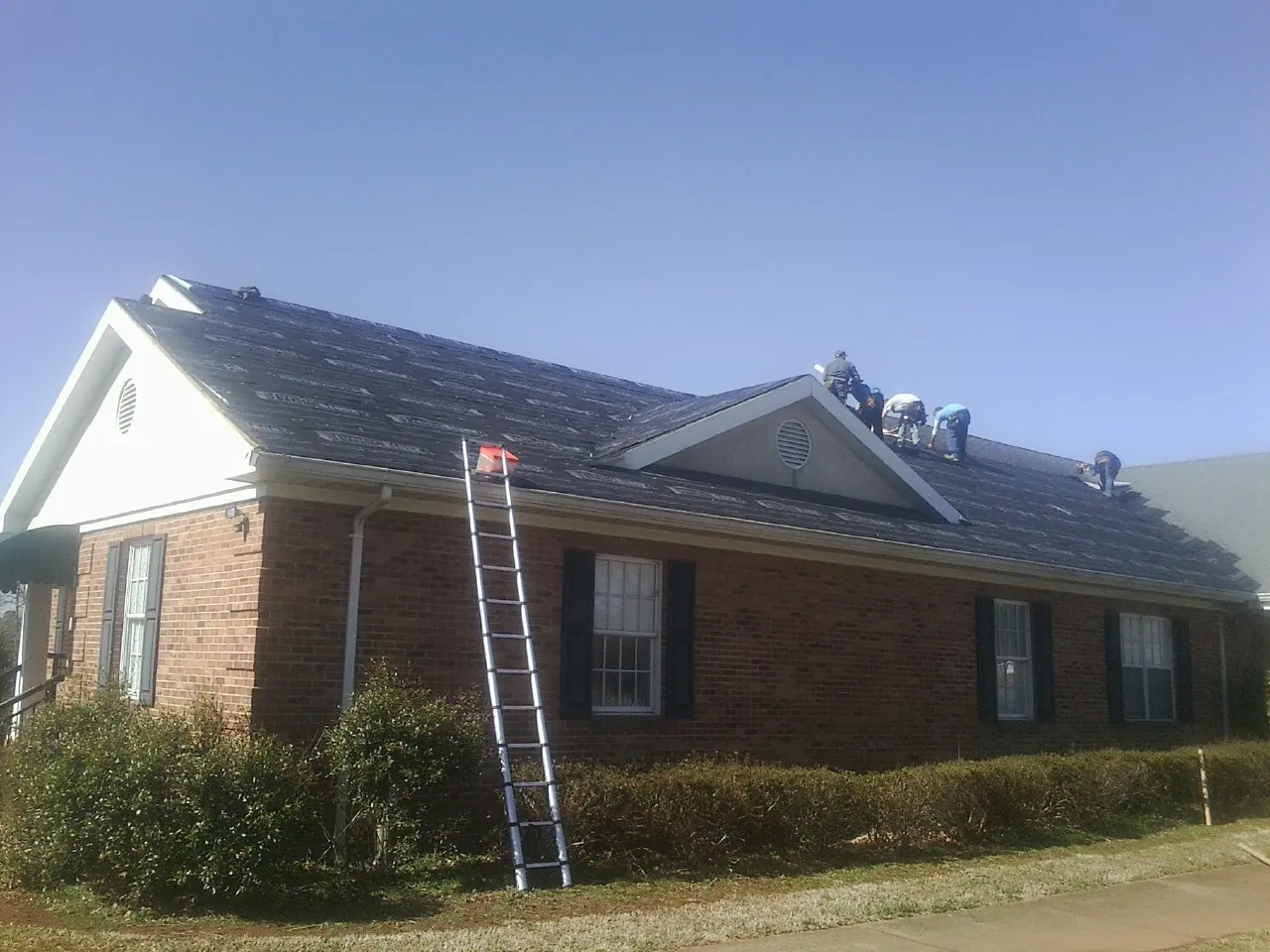 Crew tearing shingles off a brick ranch roof with a ladder set — Emergency Roof Repair work by our crew in Bristow
