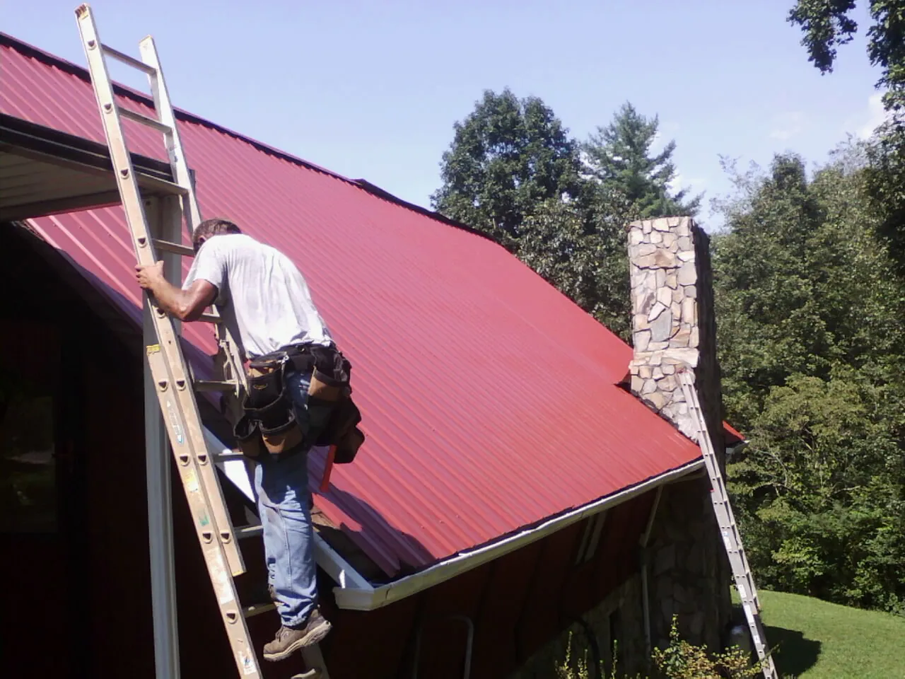 Roofer on a ladder beside a red metal roof and stone chimney — Soffit & Fascia Repair by Roofing Brothers in Bristow